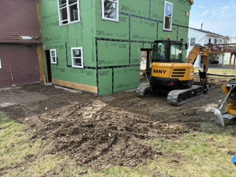 A rear yard opened up beside a green-sheathed addition, a compact excavator parked on the soil