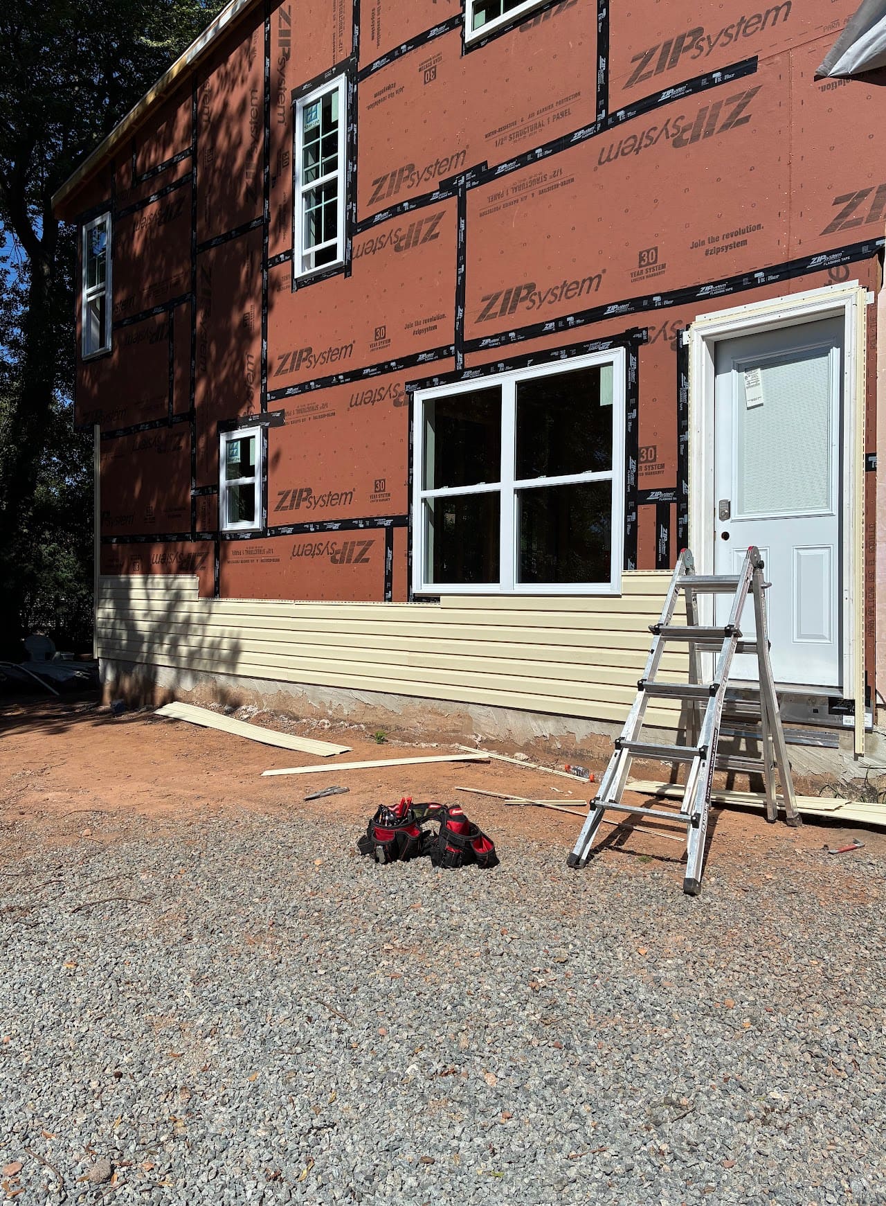 New windows and door set into brown structural sheathing, with a ladder and work boots on gravel